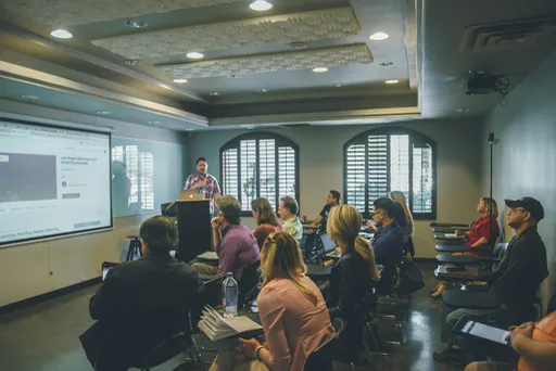 A speaker presenting to a seated room, the audience following on their laptops